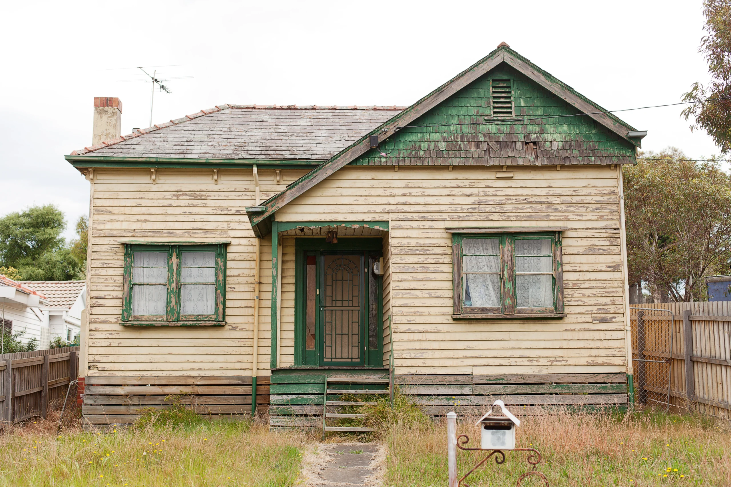 Old yellow house with peeling paint, rotted wood siding, and overgrown front yard in need of major repairs